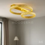 Luminaria Annelie Gold instalada en un ambiente cálido con decoración moderna. Su diseño suspendido de doble aro dorado añade lujo y personalidad al espacio, iluminando de forma suave y uniforme.