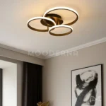 Luminaria Selene Black instalada en una sala moderna con tonos cálidos. Su diseño de aros entrelazados agrega dinamismo visual y una iluminación uniforme gracias a su potencia de 60W y 6500 lúmenes.